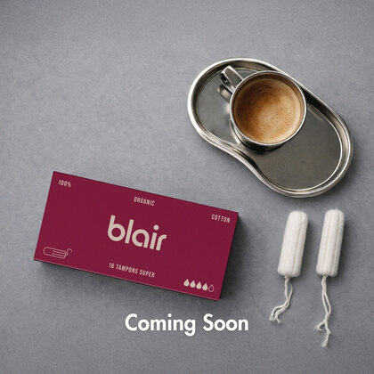 Ett vinrött Blair-paket med tamponger super bredvid en kaffekopp och två lösa tamponger på ett grått bord