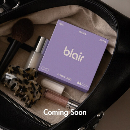 Lila Blair-förpackning med trosskydd i en väska tillsammans med sminkborste, hårklämma, synlig vit tampong och läppstift.