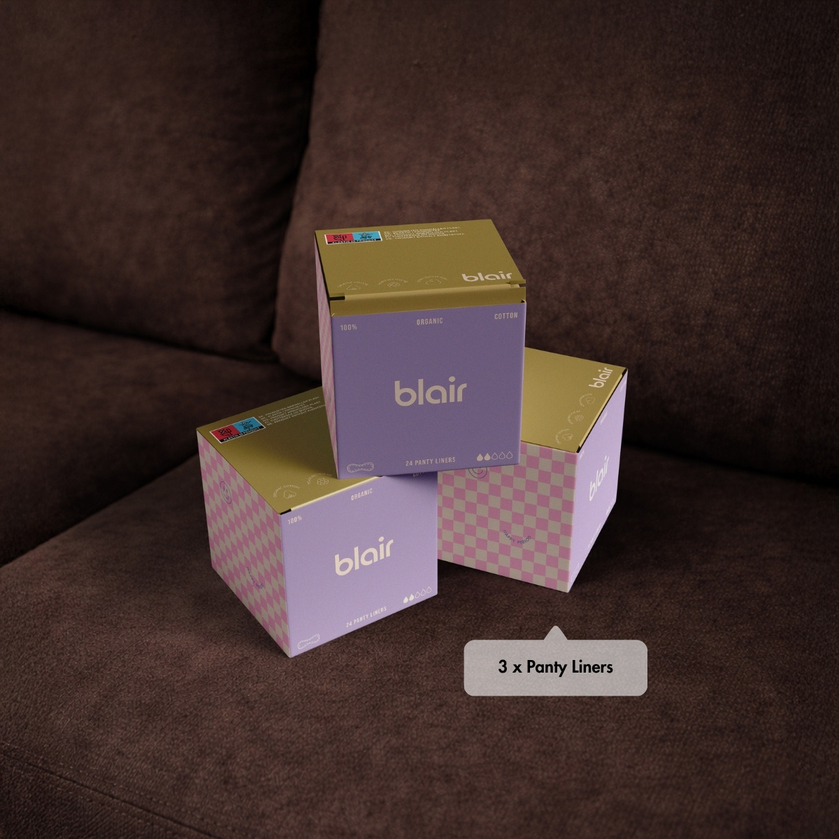 Tre förpackningar med Blair trosskydd, i rosa och lila rutigt mönster på en brun soffa, staplade på varandra