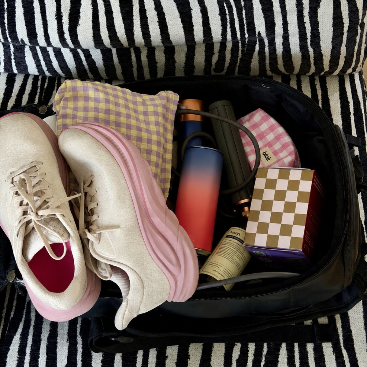En träningsväska på en fåtölj i zebra med vita löparskor, ett rutigt paket med Blair bindor och en necessär i rosa rutor