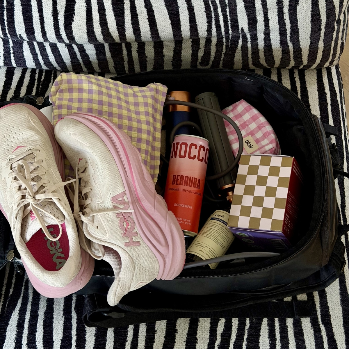 En träningsväska på en fåtölj i zebra med vita löparskor, ett rutigt paket med Blair bindor och en necessär i rosa rutor